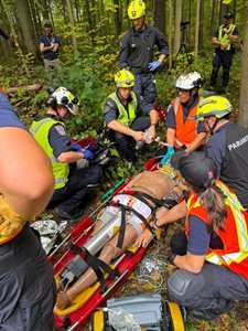 Paramedics performing a simulated medical procedure on a training mannequin secured to a stretcher in a forested area.